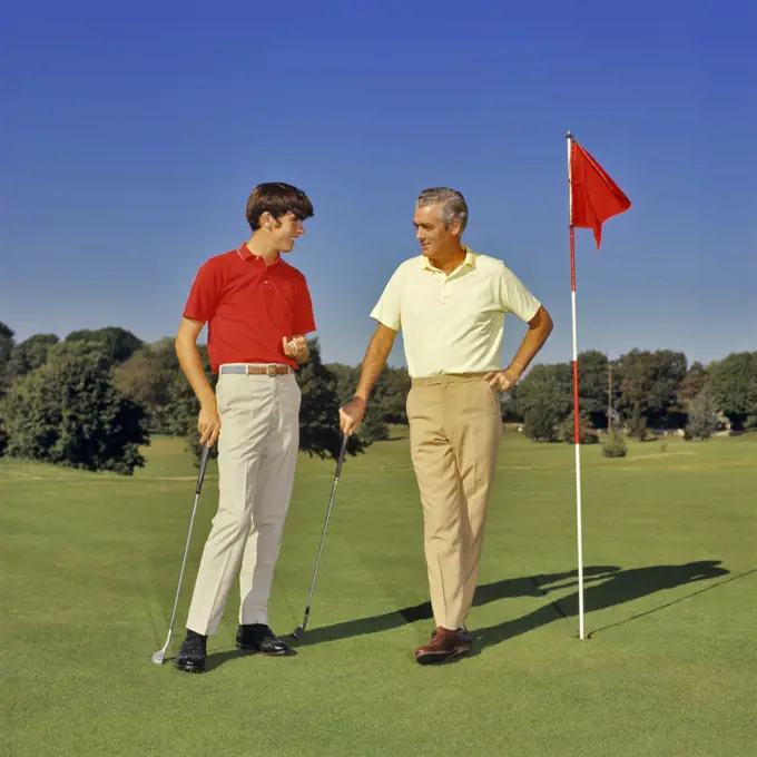1960s 1970s TEENAGE BOY AND MIDDLEAGED FATHER ON GOLF COURSE GREEN LEANING ON CLUBS TALKING