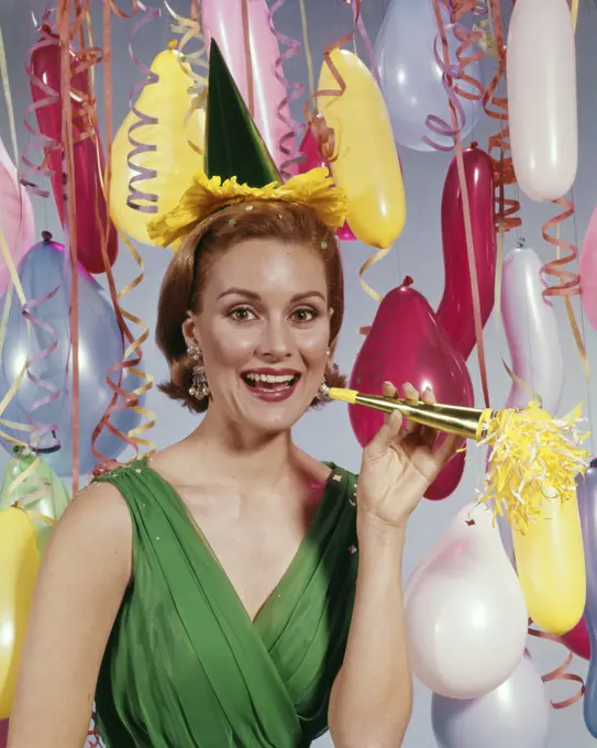 1960s SMILING WOMAN LOOKING AT CAMERA AMID STREAMERS AND BALLOONS HOLDING NEW YEARS NOISE MAKER