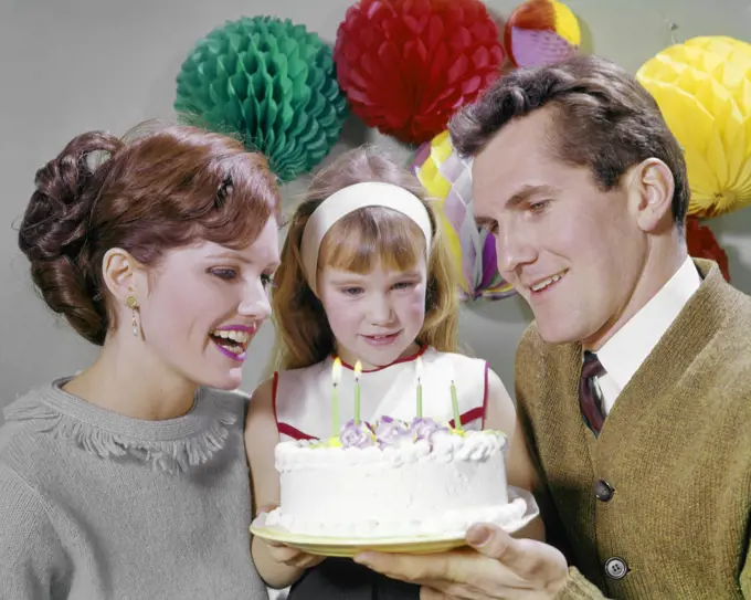1960s SMILING MOTHER AND FATHER WITH DAUGHTER READY TO BLOW OUT CANDLES ON BIRTHDAY CAKE PARTY DECORATIONS