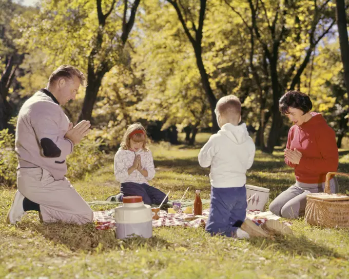 1960s FAMILY KNEELING AROUND PICNIC IN GRASS AUTUMN DAY SAYING GRACE PRAYER