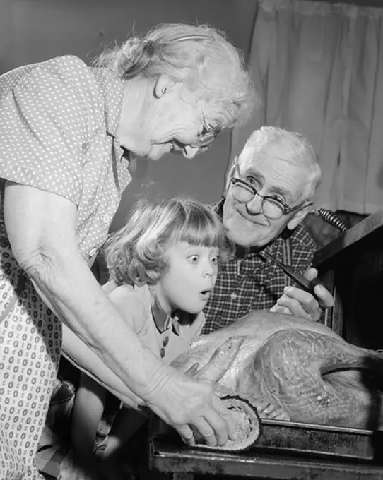 1950S 1960S Excited Little Girl Granddaughter With Grandparents Watching Cooked Turkey Coming Out Of Oven
