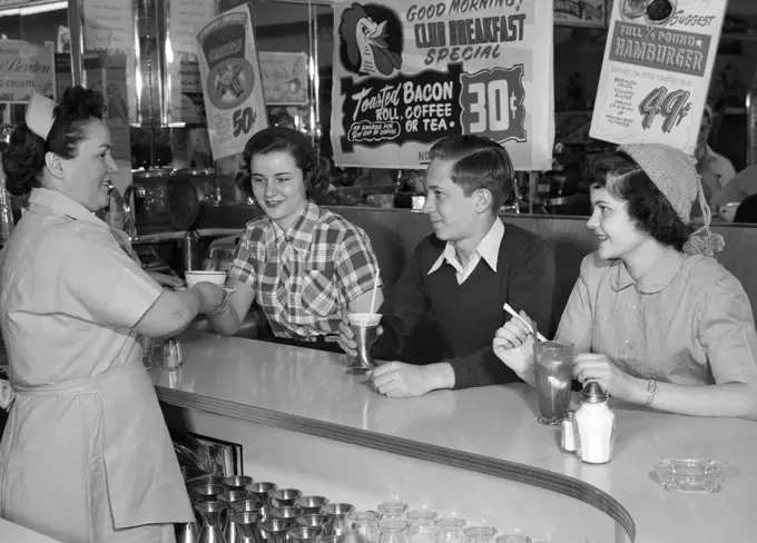 1950S Smiling Woman Waitress Serving Two Teenage Girls And One Boy At Diner Counter