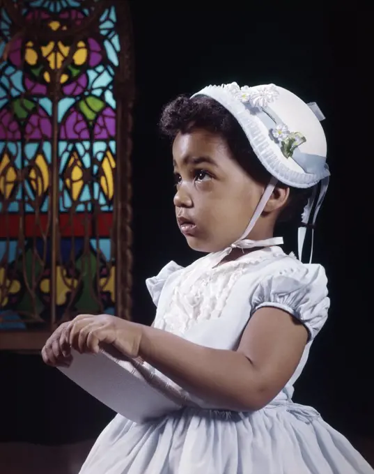 1960S Religious Little African American Girl Wearing White Hat And Dress Holding Bible Standing By Stained Glass Window In Church