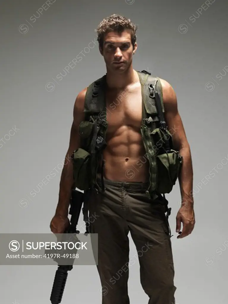 Hunky young man with a bare chest and dressed in army clothing holding an M16 automatic rifle