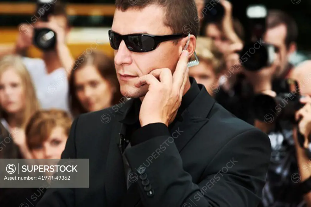 A security guard wearing dark glasses listening to his earpiece while standing in front of the paparazzi
