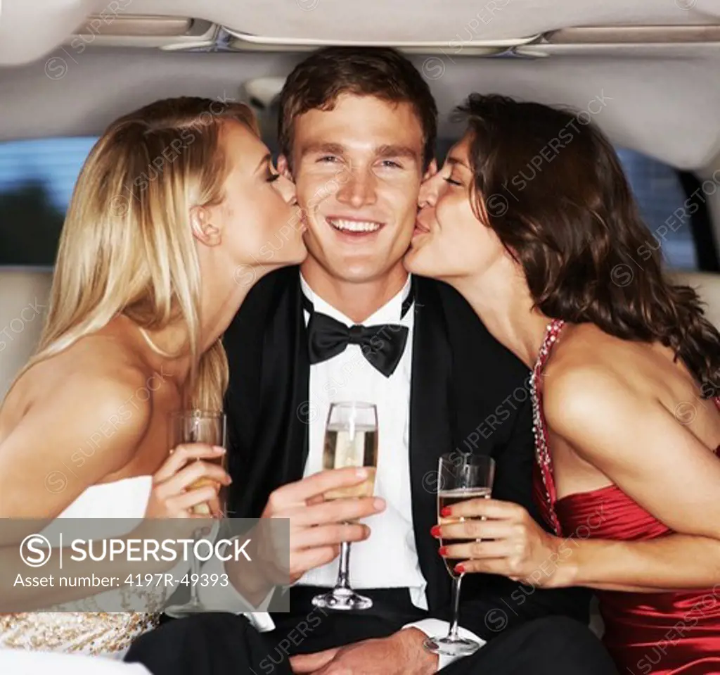 A handsome young celebrity drinking champagne and being kissed on each cheek by two gorgeous young woman