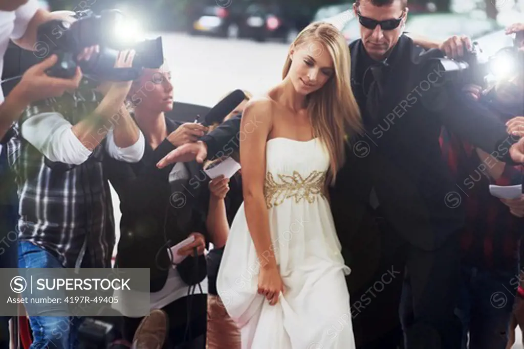 A glamorous starlet in an angelic white dress being ushered down a chaotic red carpet