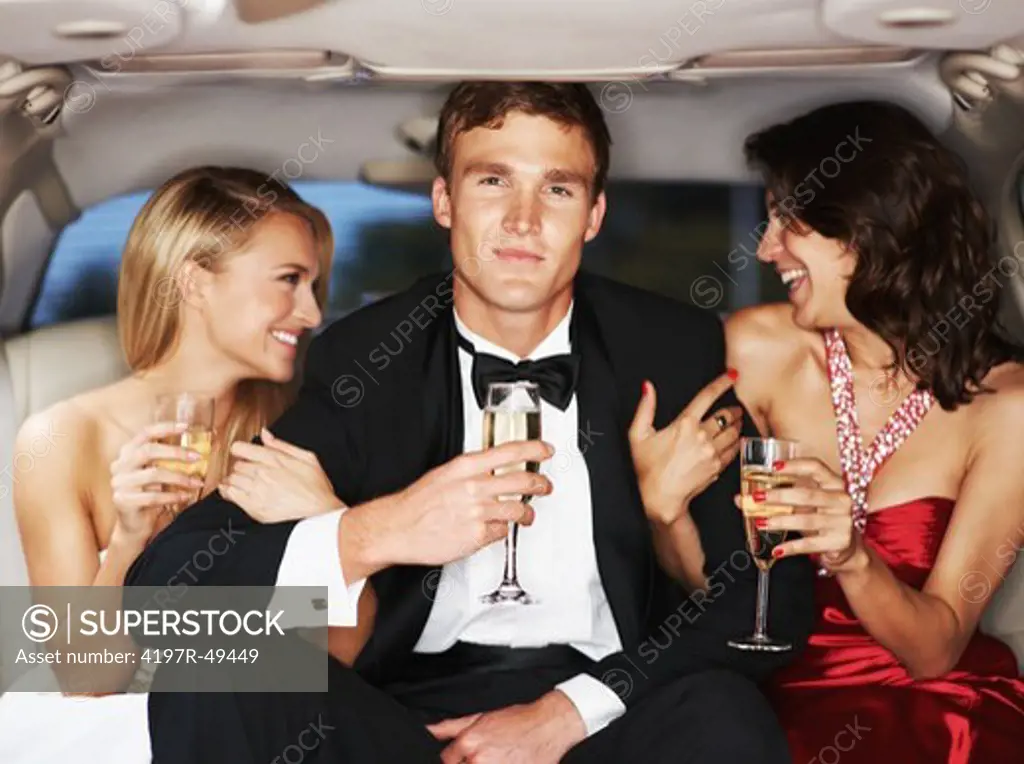 A handsome young celebrity drinking champagne flanked by two gorgeous woman in a limousine