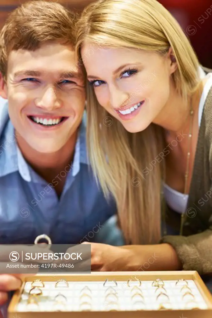 Cute young couple smiling a the camera while holding a diamond ring with a display tray in front of them