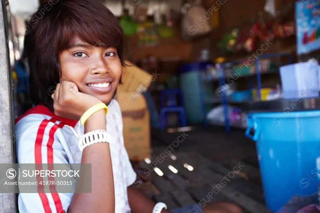 A portrait of a smiling Thai girl sitting on the ground in front of a shop