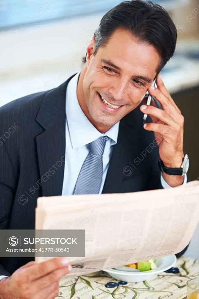 Smiling businessman using his cellphone while reading the paper over breakfast