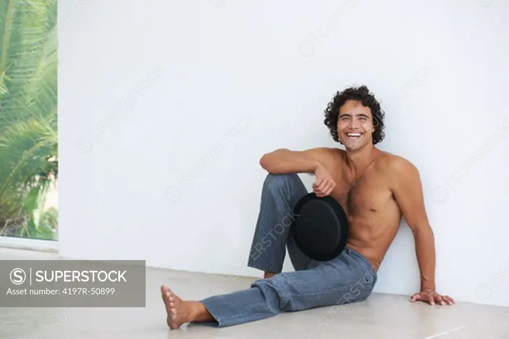 Gorgeous young man smiling at you without a shirt, holding his hat and sitting against a white wall