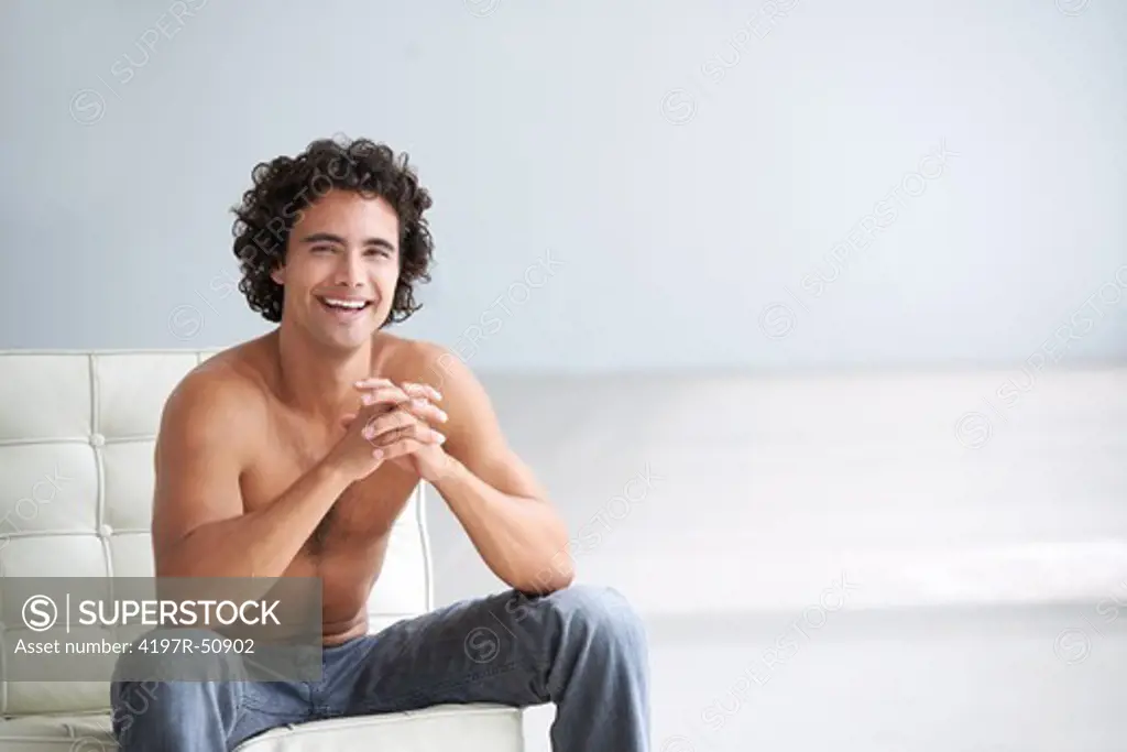 Gorgeous young man smiling at you without a shirt sitting on a chair at home