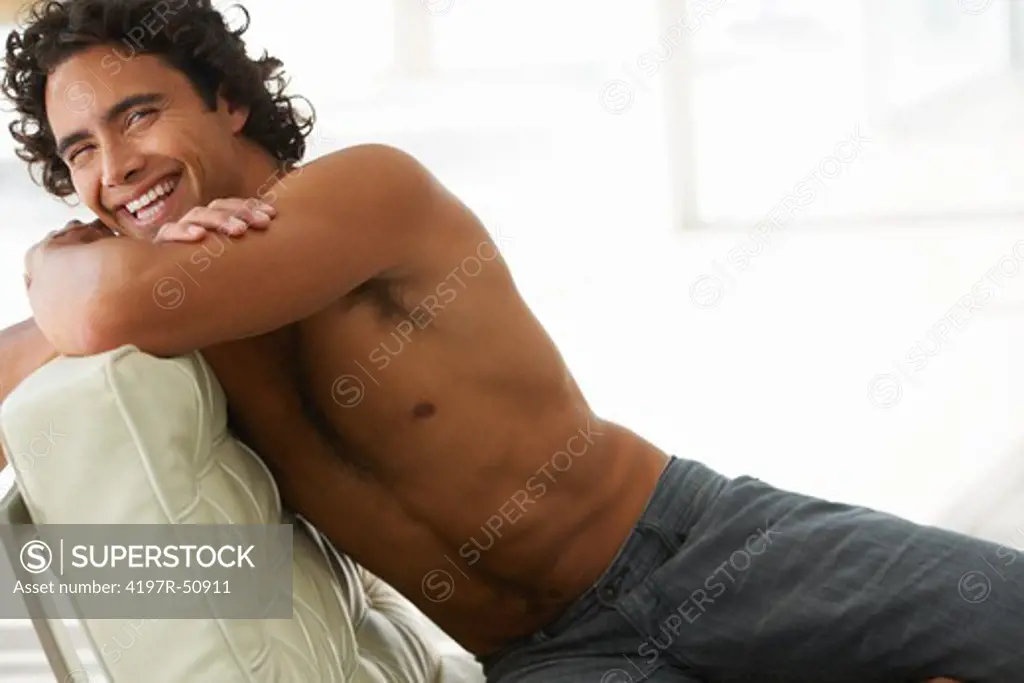 Gorgeous young man smiling at you without a shirt while reclining on  a modern sofa