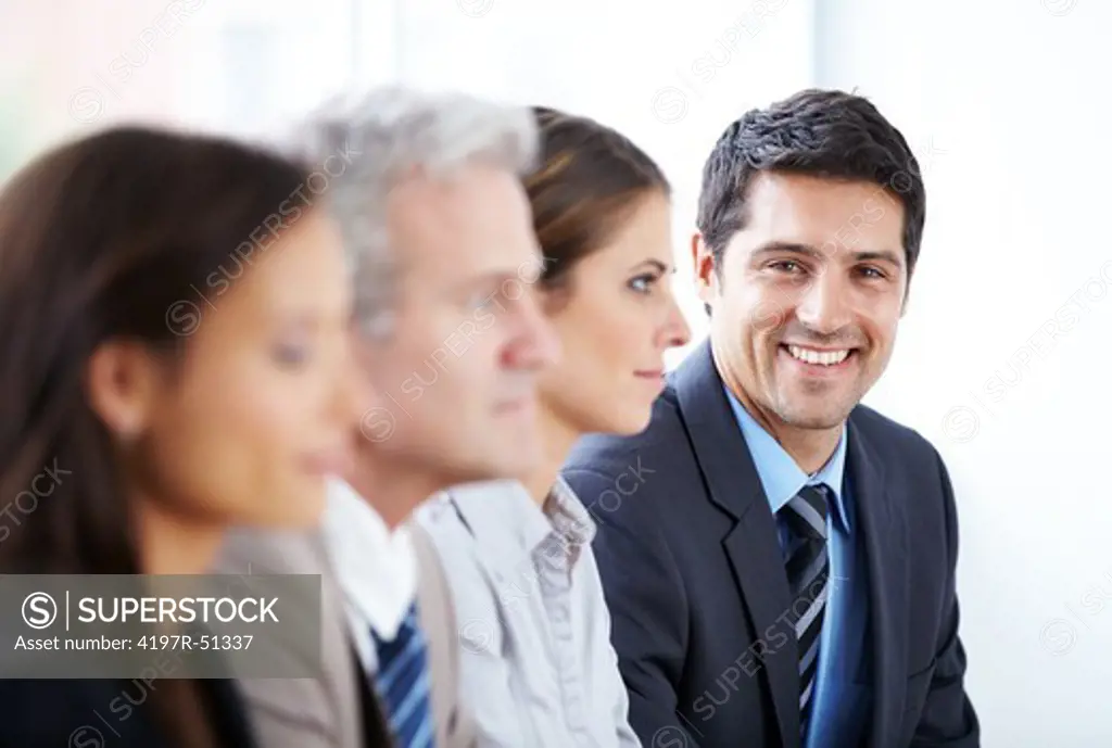 Row of businesspeople sitting at a table with the businessman at the end smiling straight at the camera