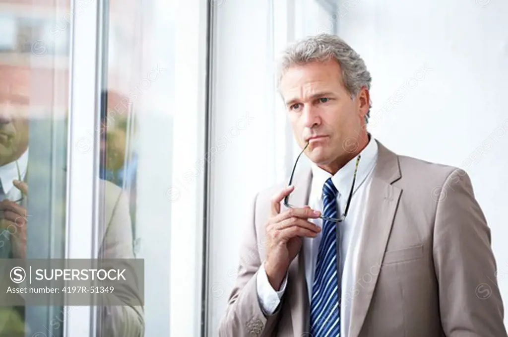 Mature businessman pensively gazing out of the window while holding his spectacles at his mouth