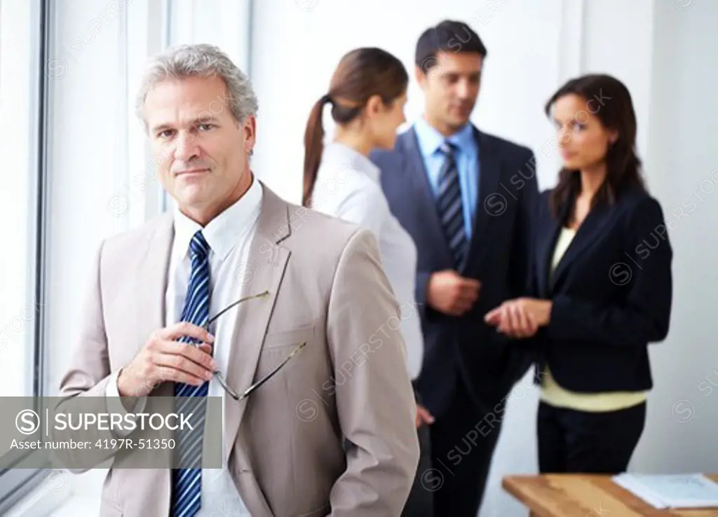 Mature businessman standing at the window smiling while his employees speak in the background