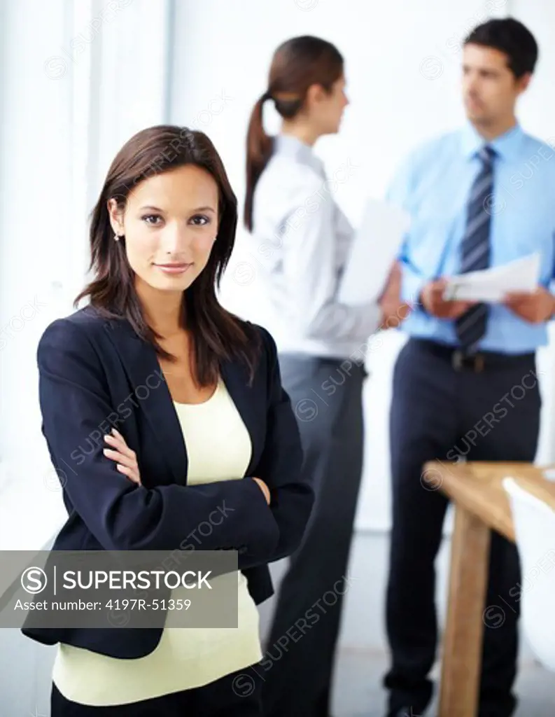 Young businesswoman standing with her arms folded and looking at the camera with two co-workers in the background