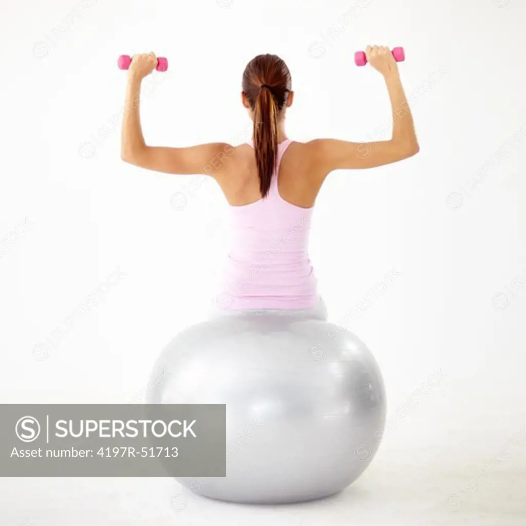 Rear view of a young woman pressing dumbbells while sitting on a gym ball - Isolated on white