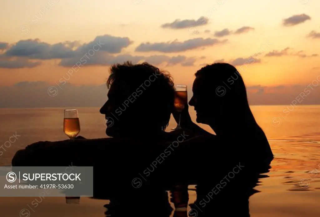 Silhouettes of a mature couple with drinks in pool by the sea in evening
