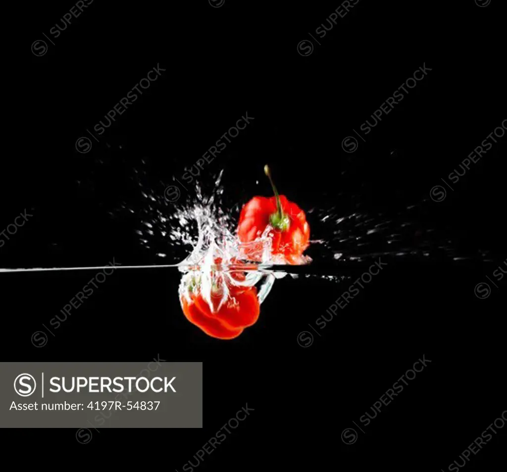 Two red peppers being dropped into water while isolated on black