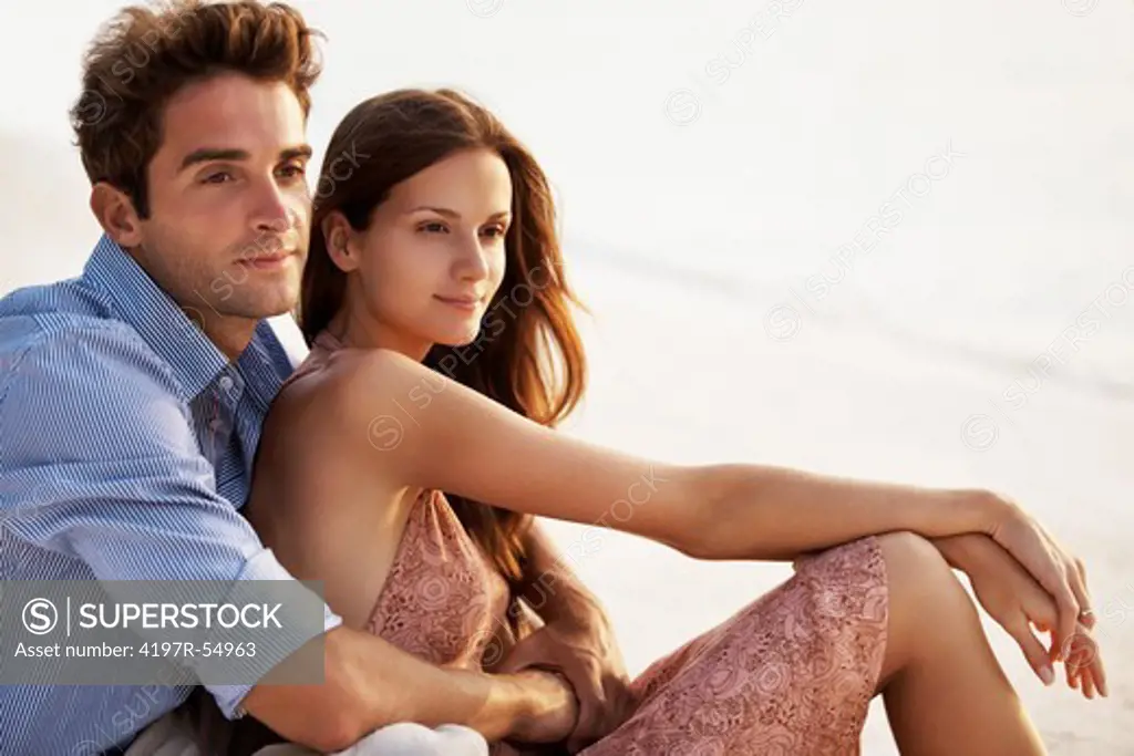 Portrait of love couple sitting close together on beach looking out at ocean