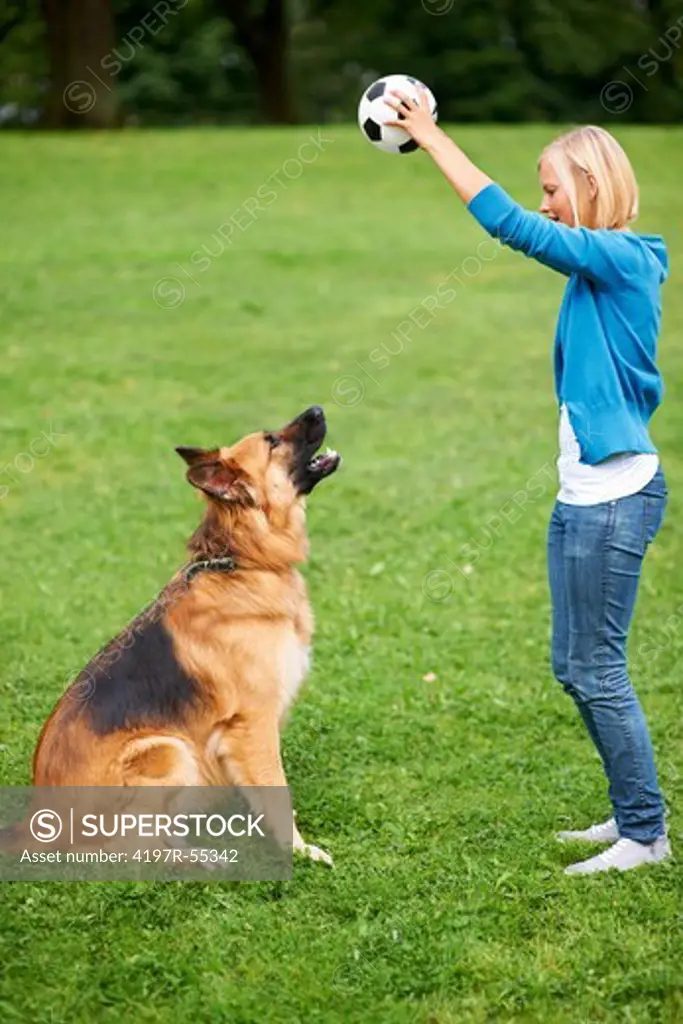 Young blonde woman holding up a soccer ball while her obedient dog sits and watches
