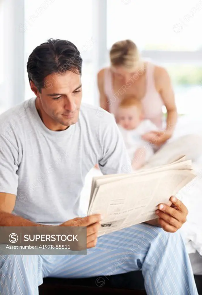 Handsome man reading the morning paper while his wife and baby play in the background