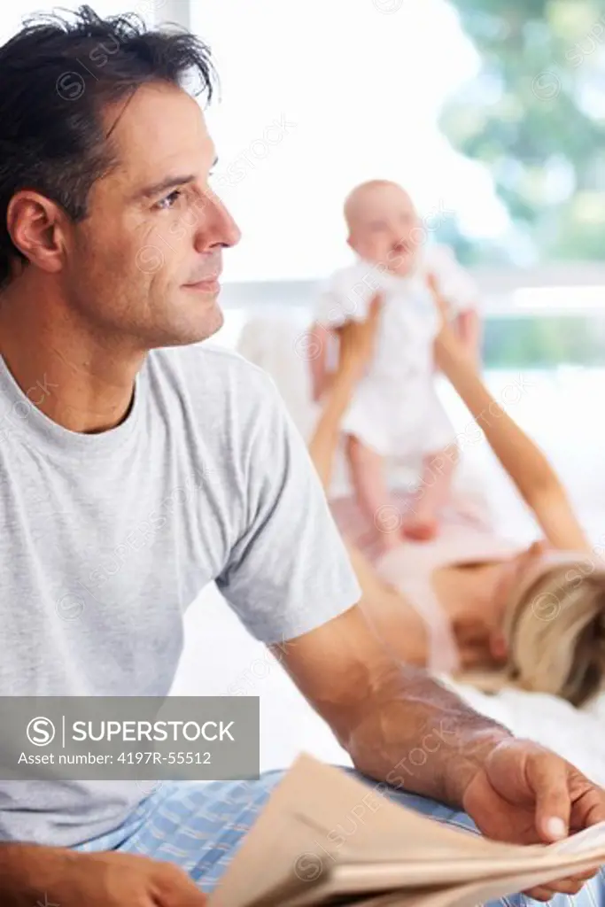 Handsome man holding the morning paper while his wife and baby play in the background