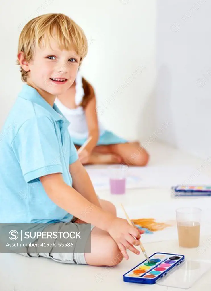 A portrait of young boy painting pictures with watercolours with his sister in the background