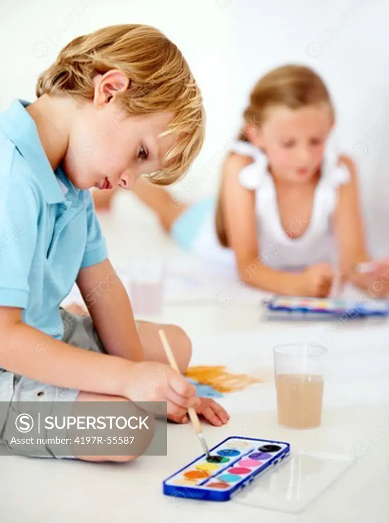 A cute young boy mixes some watercolours for his painting while his sister works behind him