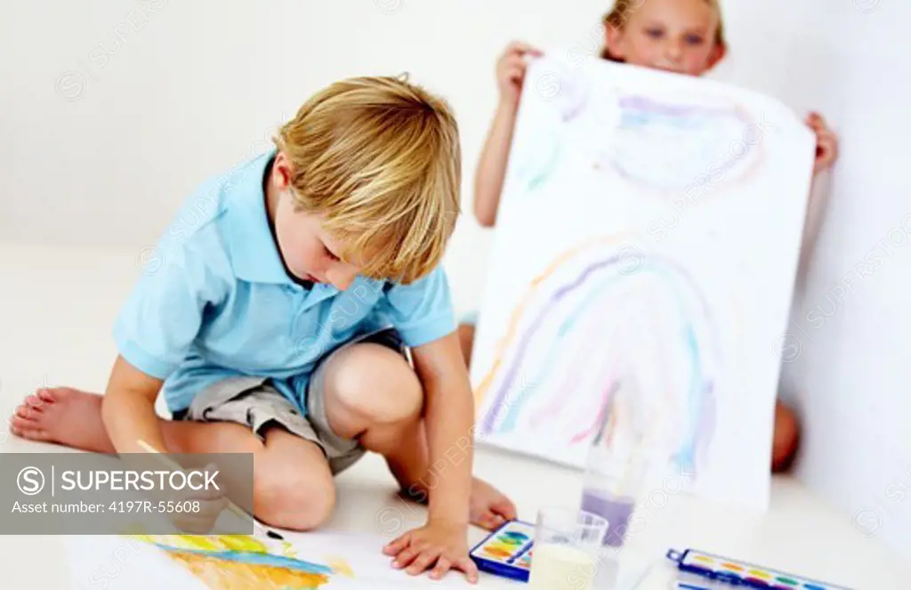 A young girl displays her artwork in the background while her brother continues working on his painting