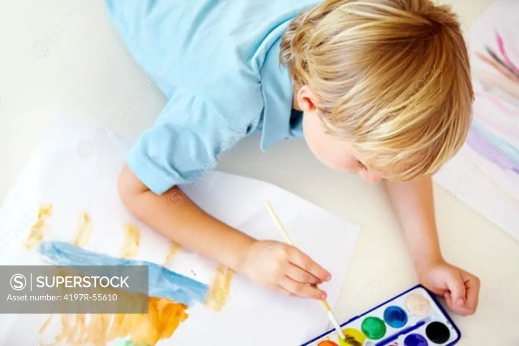 A high angle shot of a young boy painting pictures while lying on the floor