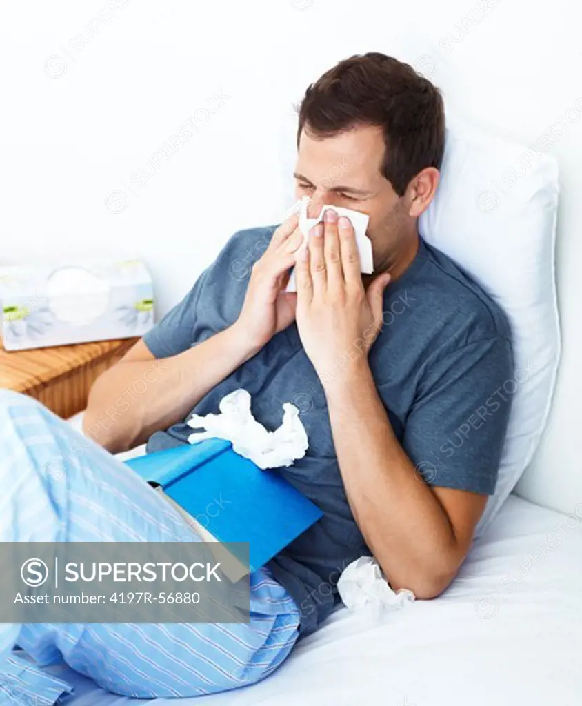 Young man lying in bed and blowing his nose while struck down with flu