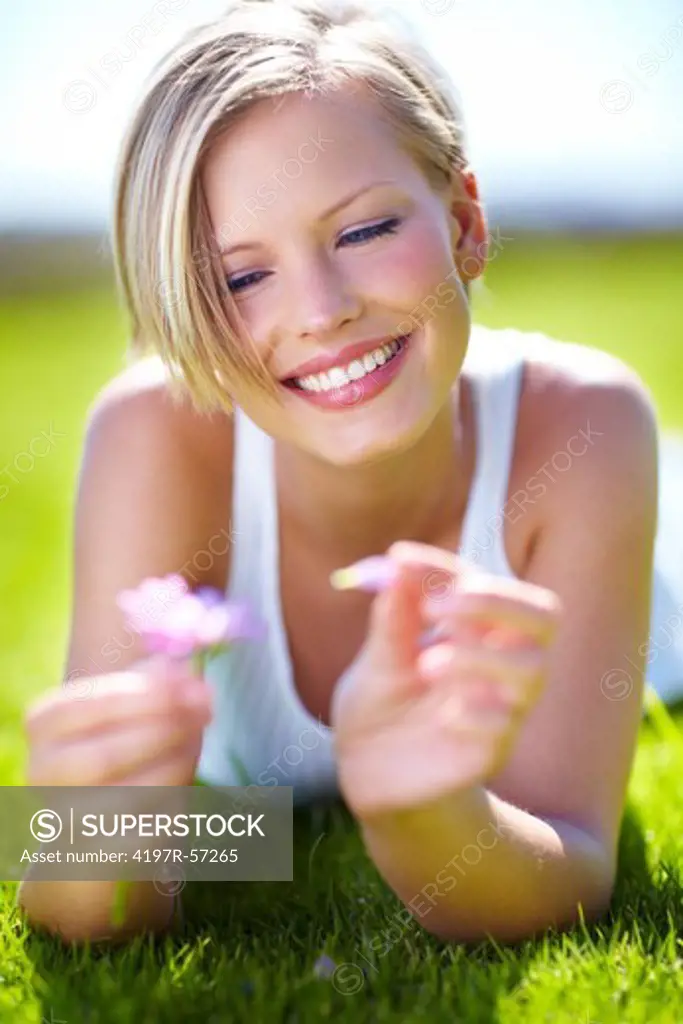 A beautiful young blonde pulling a petal off of a flower while lying on the grass