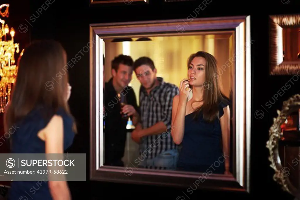 A young girl applying lipstick in a mirror while two guys check her out at the club
