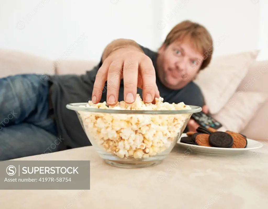 A mature man lying on the couch reaching for popcorn on the table in front of him