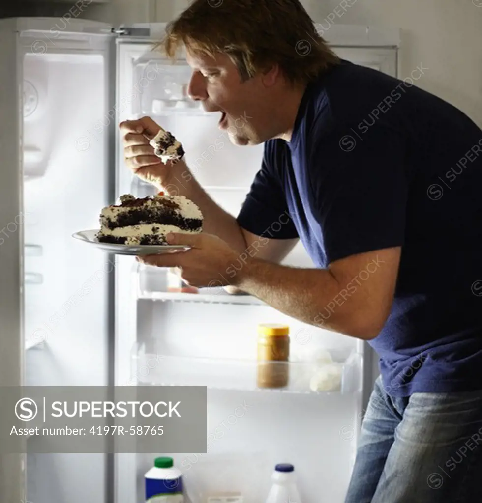A mature man in front of his fridge eating cake
