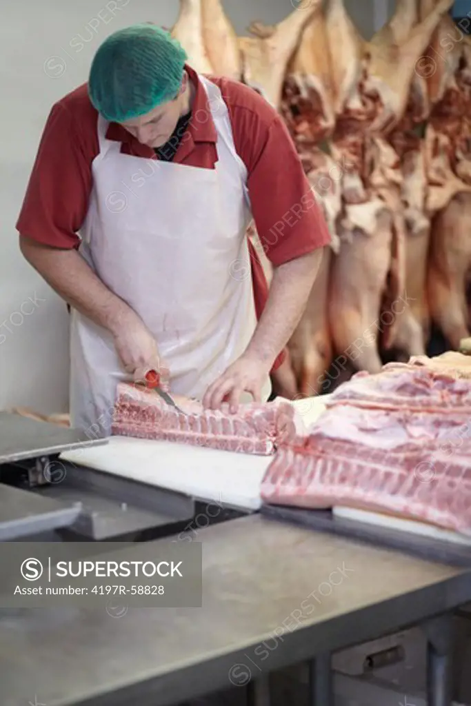 Young caucasian butcher cutiing meat with carcasses hanging in the background
