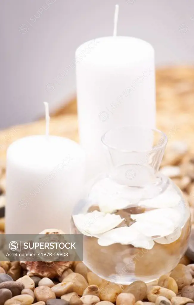 A spa arrangement consisting of candles, a seashell and a clear water jug on a bed of pebbles