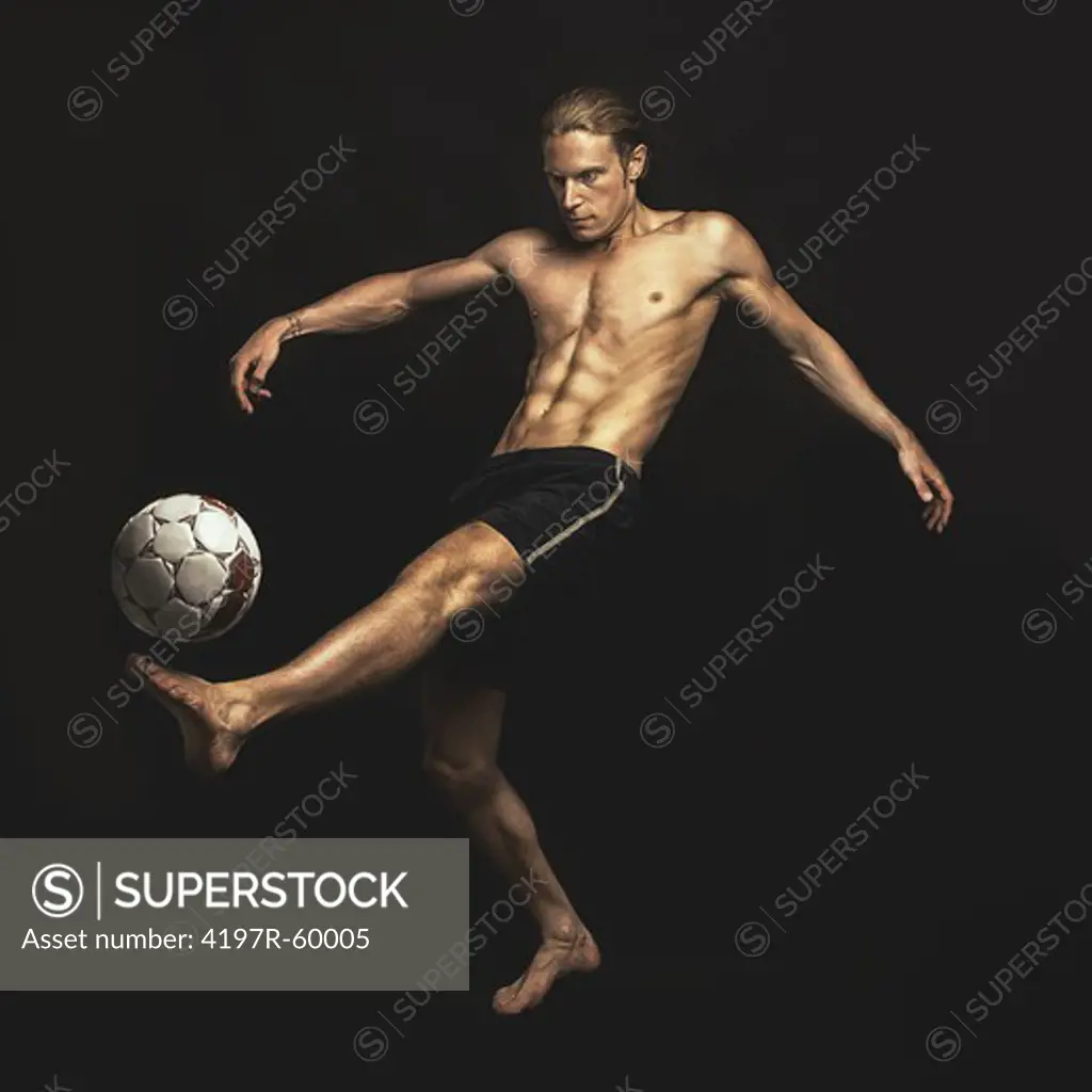 A handsome young shirtless man kicking a soccer ball on a black background