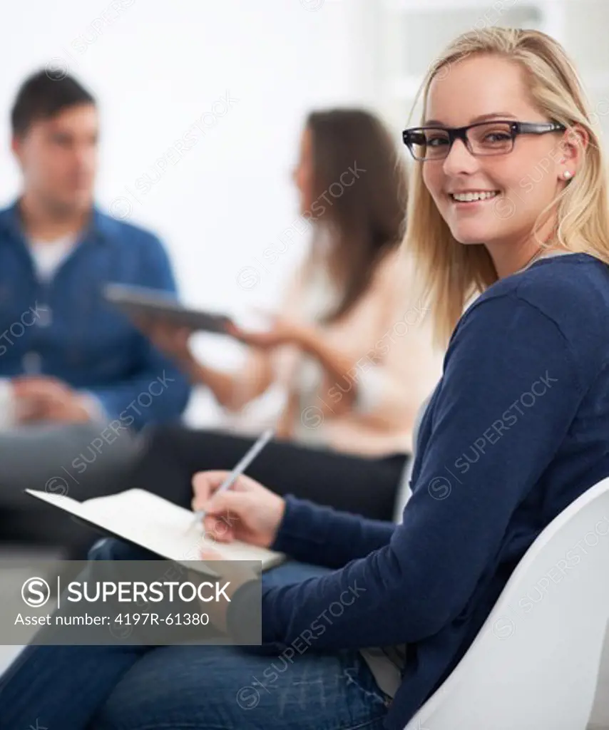 Friendly young blonde wearing glasses writing in a notebook while her colleaugues sit in the background - portrait