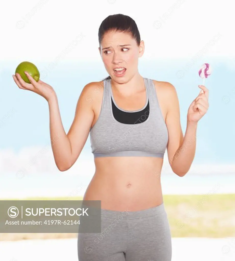 An attractive young woman looking conflicted as she decides between an apple and some candy