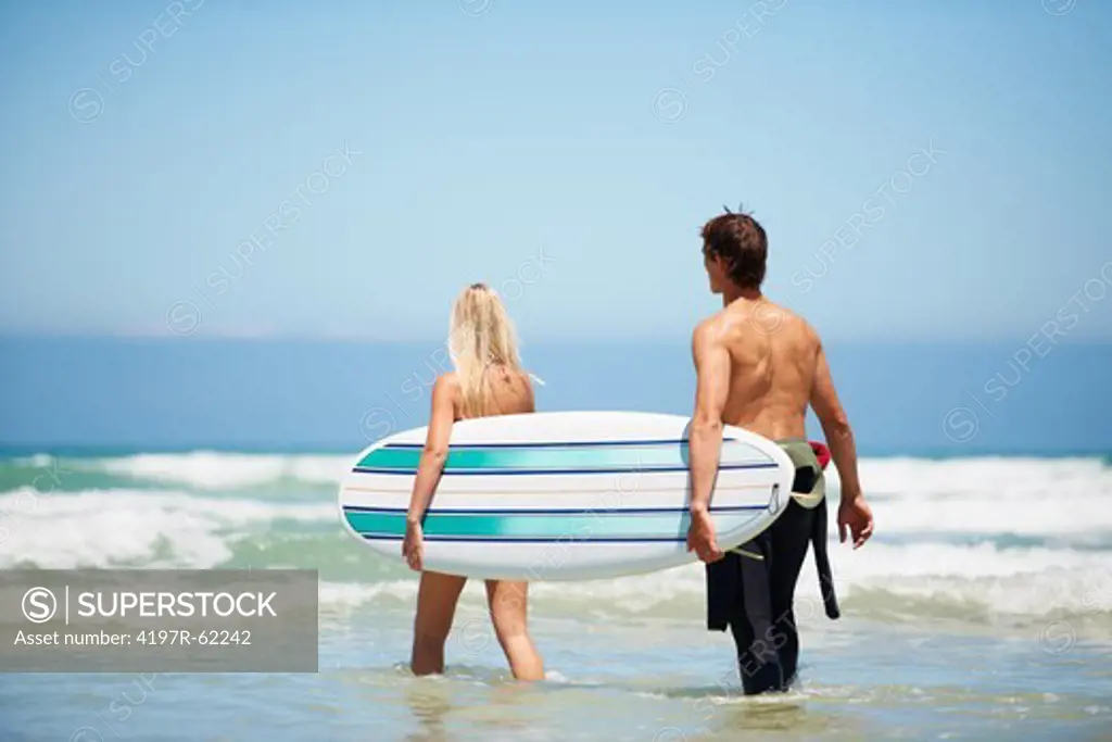 Reaview of a man and woman walking through the waves carrying a surfboard