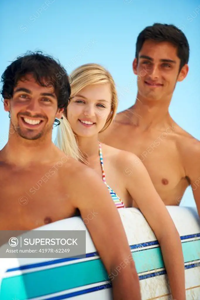 Three attractive surfers standing together holding a surfboard