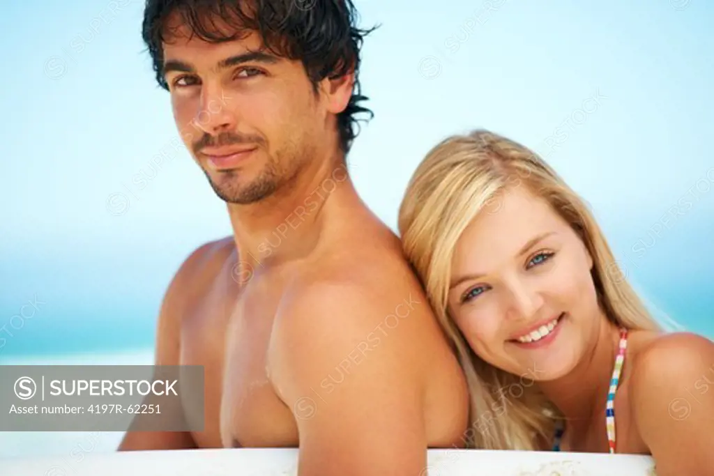 A handsome young man standing with a surfboard under his arm and his girlfriend leaning on his back