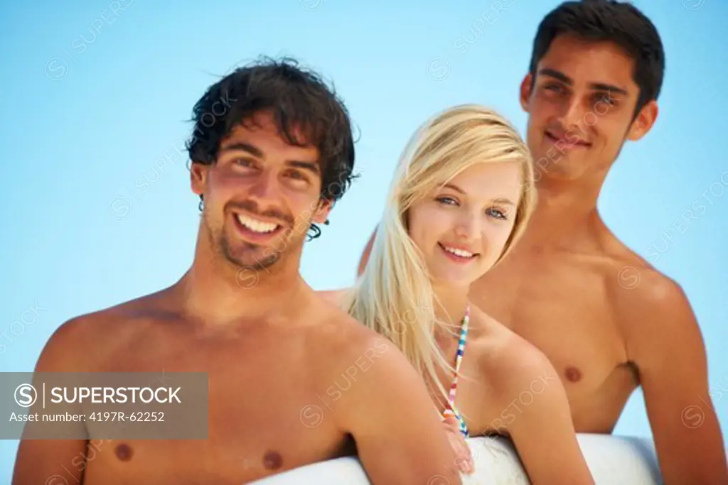 Three attractive surfers standing together and holding a surfboard under their arms