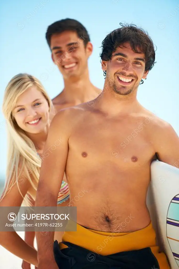 Portrait of a smiling handsome surfer standing with surfboard under his arm and two smiling friends standing behind him