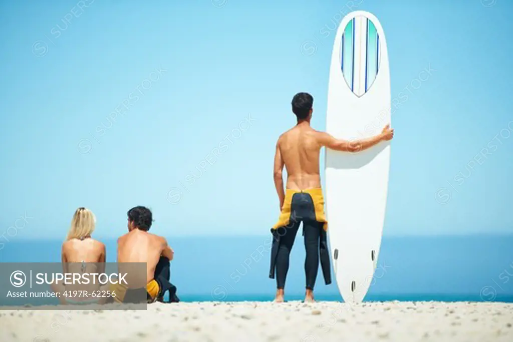 Rear view of a surfer couple sitting on a beach with a surfer friend standing besides them holding a surfboard