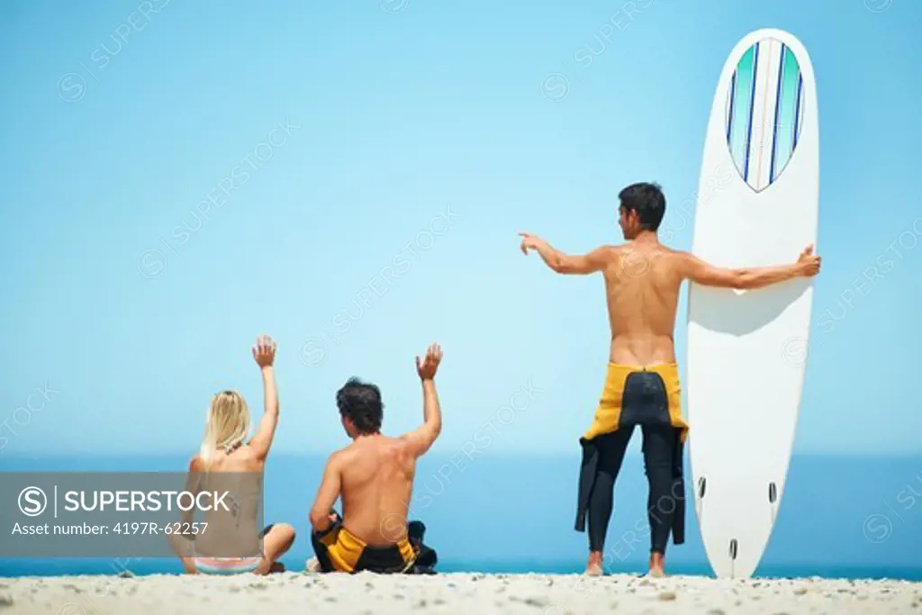 Rear view of a surfer couple sitting on a beach and waving with a surfer friend standing besides them holding a surfboard and pointing out to sea